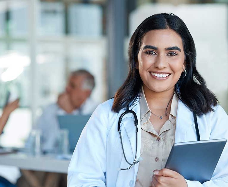 Physician Assistant smiling while holding a tablet in a hospital with other healthcare professionals