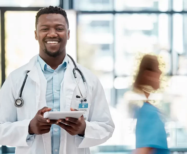 Locum tenens physician standing in a busy hospital hallway while holding a tablet and smiling